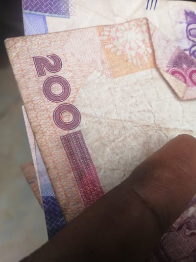 The image shows a close-up of Nigerian currency, specifically a 200 Naira note, being held by a person's finger. The note has a faint pinkish-orange background with intricate patterns. The number "200" is prominently displayed in a darker shade, with two concentric circles within the "0"s. A portion of another banknote, with purple and blue patterns, is visible behind the 200 Naira note. The scene is lit from above and slightly to the left, casting subtle shadows. The image is a still photograph taken at ground level, likely in Ajasse Ipo, Nigeria, given the context. No other elements of the environment or city are visible.