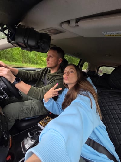 This image, captured within a vehicle in Valdgeym, Russia, displays a male driver and a female front passenger. The male, seated on the left, wears an olive green hooded sweatshirt with a "Puma" logo and olive green cargo pants, along with a dark blue smartwatch on his left wrist. His hands are positioned on the black steering wheel, and his gaze is directed forward. Above the dashboard, a black dashboard camera or recording device is mounted. The female, seated in the front passenger seat and angled towards the camera, wears a light blue hooded sweatshirt. She is pouting her lips and making a "peace" sign with her left hand, which rests on the driver's left forearm. Her fingernails are painted in multiple vibrant colors. A black seatbelt is visible across her chest. The vehicle's interior features black, presumably quilted, seat upholstery. The center console includes an automatic gear shifter and a silver electronic parking brake button. A notebook or organizer with visible text, including "ACHTUNG," is placed next to the gear shifter. Through the vehicle's windows, dense green foliage and trees are visible, indicating a natural or wooded outdoor environment.
