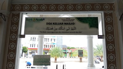 This is a medium shot, captured from inside a building, looking outwards through a large doorway. The doorway is framed by decorative pillars and a patterned frieze. A large banner is prominently displayed across the doorway. The banner features the Bank Aceh logo and text in both Indonesian and Arabic. The Indonesian text reads: "DOA KELUAR MASJID" (Prayer for Leaving the Mosque), and below it, a translation: "Ya Allah, sesungguhnya aku memohon karunia dari-Mu" (O Allah, indeed I ask for Your favor). Above this, the banner also indicates: "MASJID AGUNG SULTAN JEUMPA KABUPATEN BIREUEN".
Outside the doorway, a street scene is visible. The architecture suggests a warm climate, with palm trees and other vegetation. There are several buildings, including what appears to be a shop or commercial establishment with red awnings. A few people are present. One person wearing a blue shirt is seen bending down on the left side of the frame. Further back, a person is riding a motorcycle with a passenger, and another person appears to be standing or walking on the right. A QR code is affixed to a pole on the right side of the scene. The overall impression is a view from a building with religious or public significance in a bustling Indonesian urban or semi-urban environment.