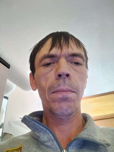 The image is a close-up, head-and-shoulders selfie of a Caucasian male in what appears to be an indoor setting. The man has medium-toned skin, thinning dark brown hair, and is looking directly at the camera with a neutral expression. His face shows signs of aging, with wrinkles around the eyes and mouth. He is wearing a gray zip-up jacket with a collar. The background is out of focus, but suggests a domestic environment with a light-colored ceiling and some wooden cabinetry visible on the right side. There are no explicit indicators within the image to suggest a specific location in the city of Zyuzino, Russia.