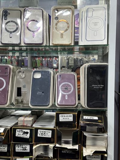 The image shows a retail display of phone cases in Bireuenmeunasahreuleut, Indonesia. The display is organized on multiple shelves within a glass-fronted cabinet. The top shelves feature various phone cases in their packaging, many designed for specific phone models such as "For SM A15 4G/5G," "A54 5G," and "For SAM A17 Clear Case." Some cases are transparent with magnetic rings, while others are colored silicone.
The middle shelves continue the display of phone cases, with some for "iPhone 15 Silicone Case" and "iPhone X/XS Silicone Case." A small glass bottle, possibly containing a cleaning solution or accessory, is placed next to one of the iPhone cases. The bottom shelves showcase stacks of boxes, some labeled with product names and quantities, such as "FOR SA A24 4G" and "25PCS/BLACK." The boxes appear to contain more phone cases or related accessories.
The overall environment suggests a mobile phone accessory shop. The lighting is adequate, highlighting the products on display. No people are prominently featured in the image, though a partial reflection of a person is visible in the glass cabinet on the middle shelf. The arrangement of items is neat and organized, indicating a typical retail setting.