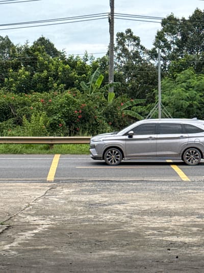 The image shows a silver SUV driving on a two-lane road in Phú Quý 2, Vietnam. The car is in the right lane, with a yellow line marking the lane division. To the left of the road, there is a guardrail and lush green vegetation, including trees and bushes with red flowers. In the background, there are power lines and utility poles. The sky is overcast. The foreground shows a concrete surface with some cracks and water marks.