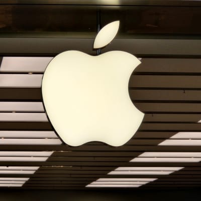A close-up shot features the illuminated Apple logo, a stylized apple with a bite taken out of it, mounted on a building facade. The logo is a soft, creamy white and appears to be backlit. To the left of the main logo, a smaller, leaf-shaped element of the logo is visible, also illuminated.

The background consists of vertical paneling, alternating between darker, textured surfaces and lighter, almost white surfaces that appear to be illuminated from behind. This creates a striped effect. Sunlight casts diagonal shadows across these panels, suggesting it is daytime. The building material appears modern and possibly made of metal or glass.

There is no visible text in the image. The scene is a static shot of the Apple logo, implying it is part of the exterior of an Apple store or corporate building. The overall impression is one of modern design and clean aesthetics, typical of the Apple brand. No people or specific activities are depicted. The lighting and shadows suggest a sunny day, but no information about the weather is directly available. The location context of Zaragoza, Spain, is not visually evident within the image itself.