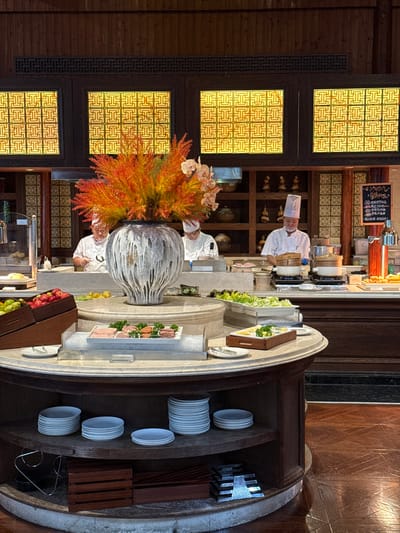 This image depicts an interior view of a buffet or open kitchen area within a restaurant in Tongxiang, China. The scene is dominated by a central, circular buffet island made of wood with a light-colored stone or marble top. Atop this island, a large, textured white and grey vase holds an arrangement of vibrant orange and red foliage, accented by several pink orchids.

On the buffet island, various food items are presented: a wooden crate holds fresh red apples and green citrus fruits; a rectangular white tray containing sliced meats (appearing as ham and salami) is displayed on a bed of ice, accompanied by serving tongs; and multiple trays of fresh green salad ingredients are visible. Below the countertop, circular shelves on the wooden island hold neatly stacked white plates of varying sizes.

In the background, behind the main buffet, an active open kitchen or serving line is visible. Three chefs, dressed in white chef coats and hats, are positioned behind a counter. One chef on the right, wearing blue gloves, is actively attending to two white pots with bamboo steamers placed on burners. Another chef is visible towards the left, behind the floral arrangement. The background features dark wood paneling and shelving units displaying decorative ceramic items. Above the kitchen area, four illuminated rectangular panels with intricate, repeating geometric patterns (possibly traditional Chinese motifs) cast a warm yellow light. The flooring is dark wood with a distinct parquet or herringbone pattern. On the far right, a vertical sign with handwritten Chinese characters and a beverage dispenser are present.