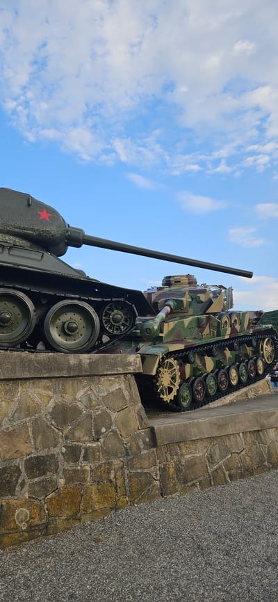 Two historical tanks are displayed on a stone embankment under a partly cloudy blue sky in Svidník, Slovakia. The tank on the left is dark green with a red star emblem on its turret and appears to be a Soviet T-34 model, characterized by its prominent road wheels and distinctive hull shape. The tank on the right is camouflaged with brown, green, and beige patterns and features a German cross emblem on its turret, suggesting it is a German Panzer IV model. Both tanks are positioned with their turrets facing towards the right of the frame. The embankment is constructed of irregular, light-colored stones. In the background, a hill is visible on the right, and the sky is filled with white, scattered clouds against a bright blue backdrop. The ground in front of the embankment is covered with small, dark gray gravel. No people are clearly visible interacting with the tanks in this frame.