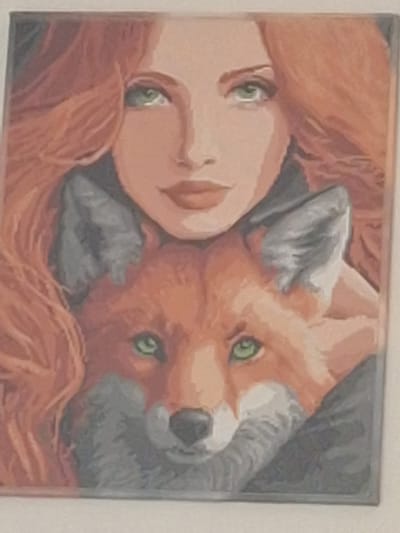 This media file depicts a painting of a person with red hair and a fox. The subjects appear to be nestled together, with the fox's head resting near the person's face. Both subjects have green eyes. The person's face is rendered in soft, warm tones, and their red hair flows around them. The fox is painted in characteristic orange and grey hues, with its ears prominent. The background is indistinct, suggesting a close-up focus on the two figures. The style of the painting appears to be representational, possibly oil or acrylic on canvas. There is no visible text in the image. The context provided indicates the location as Mińsk Mazowiecki, Poland, but there are no specific visual cues within the painting to suggest this location. The overall mood conveyed by the painting is one of intimacy and connection between the person and the fox. There is no indication of a specific time of day or weather conditions.