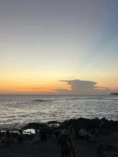 The image captures a sunset at Pererenan Beach in Indonesia. The sky transitions from a pale grey-blue to a vivid orange and yellow near the horizon. A dark cloud hovers in the center. The ocean reflects the sunset's colors, with waves crashing against a rocky shore. A group of people are gathered on the dark rocks near the water's edge, observing the sunset. Their actions suggest a moment of shared observation and appreciation of the natural environment.