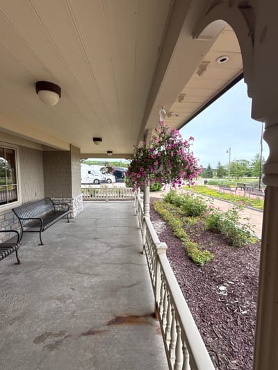 This image shows a covered porch with a concrete floor in front of a building. There are two dark metal benches on the porch. A hanging basket of pink flowers is suspended from the ceiling of the porch. In the background, there is a white semi-truck and a black semi-truck parked. Beyond the trucks, there is a landscaped area with benches and trees. The sky is overcast. The image appears to have been taken in Bemus Point, United States.