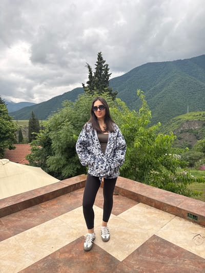 A young woman with dark hair and sunglasses stands on a tiled balcony in Akhtala, Armenia. She is wearing a black and white patterned jacket over a dark top, black leggings, and white athletic shoes. Behind her, lush green mountains rise under a cloudy sky. Trees and a red-tiled roof are visible in the middle ground, and a portion of a light-colored awning can be seen on the left side of the frame. The balcony floor is covered in large square tiles with a marbled pattern in shades of beige and brown.