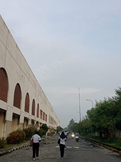 The image depicts a street scene in Sukomoro, Indonesia. The street is flanked by a long, low-rise building on the left, characterized by repetitive arched openings filled with what appears to be a patterned screen. On the right, a row of trees lines the street.
Two individuals are walking away from the viewer, dressed in what appears to be uniform-like clothing. Further down the road, there are more people and motorbikes with headlights on. Streetlights are visible intermittently along the roadside. The sky is overcast and the road surface appears damp. The general environment suggests an early morning or late afternoon setting.