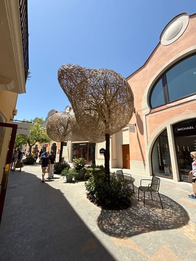 The image captures an outdoor pathway in what appears to be a shopping outlet or village, specifically identified as La Roca Village in La Roca del Vallès, Spain. The architecture features buildings with peach-colored facades and arched windows. In the foreground, two large, spherical woven art installations resembling trees are prominent. A narrow pathway, paved with large grey tiles, leads into the distance, lined with storefronts. Several people are walking along the pathway, some carrying shopping bags. The scene is bathed in bright sunlight, casting distinct shadows from the art installations and the surrounding trees onto the ground. Two modern, dark metal chairs are placed near the art installation on the right side of the frame. Store signs visible include "BROWNIE," "GANT," and "MICHAEL KORS." The sky is a clear, bright blue.
