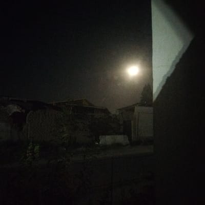 The image depicts a night scene with a bright, full moon illuminating the sky above a few structures in Akkurgan, Uzbekistan. In the foreground, a pale wall casts a shadow that occupies the right side of the frame. Behind this wall, some vegetation and the roofs of low-rise buildings are visible, appearing dark and silhouetted against the moonlit sky. The overall impression is that of a quiet, moonlit night in a residential area.