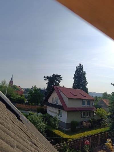 The image shows a view of the city of Niemcza, Poland, on a clear day. In the foreground, a tiled roof is visible, partially obscuring the view. Beyond the roof, a two-story house with a red-tiled roof is prominent, featuring white window frames and a light green facade. A satellite dish is mounted on the side of the house. In the background, a church with a tall, dark spire can be seen, along with other buildings and lush green trees. The sky is a pale blue with a few wispy clouds. Two small, dark objects, possibly birds or distant kites, are visible in the upper left portion of the sky. The overall scene depicts a residential area in a European town.