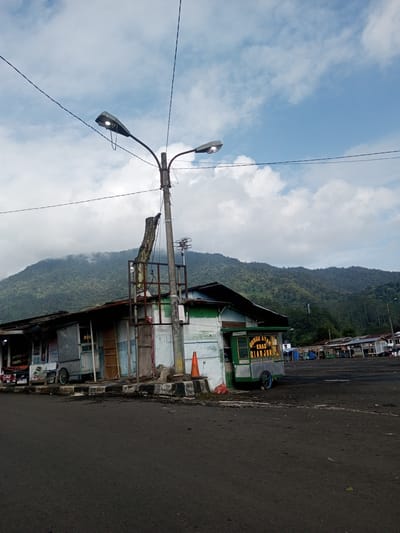 The image shows a landscape in Mya Sit Kaing Ywar Thit, Myanmar featuring a street scene with a building complex in the foreground against a backdrop of a mountain range. The buildings appear to be weathered and somewhat dilapidated, with corrugated metal roofing and faded paint. A street light pole is positioned near the buildings, with visible power lines extending into the sky. The area in front of the buildings is paved and shows signs of wear. A food stall cart is on the side of the buildings. The sky is partly cloudy, with the mountain range extending across the horizon, partially obscured by clouds. The mountain is covered in lush, green vegetation. The scene suggests a rural or semi-rural environment.