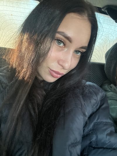 A young woman with long, dark hair and light eyes is lying down, possibly in a car. She is wearing a dark, puffy jacket. The background suggests a window with horizontal lines, potentially a defroster grid. The location is unspecified, and there is no evidence to confirm it is Kvarsa, Russia.