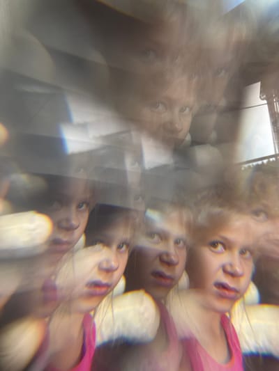 The image shows a distorted and fragmented view of several young children's faces, likely siblings, due to a lens effect or multiple exposures. Their expressions appear to be a mix of surprise, curiosity, and slight discomfort. The children are close together, suggesting an indoor setting, possibly a home or a studio. The bright lights and colors, particularly the pink top worn by one of the children, are prominent, adding to the overall abstract and dreamlike quality of the photograph. The children's features, such as their eyes and mouths, are clearly visible despite the distortions. The background is blurred and indistinct, with hints of an interior space and perhaps an outdoor scene visible through a window or glass. The image does not provide any specific clues about its location in Baryshevo, Russia, or any other geographical context.