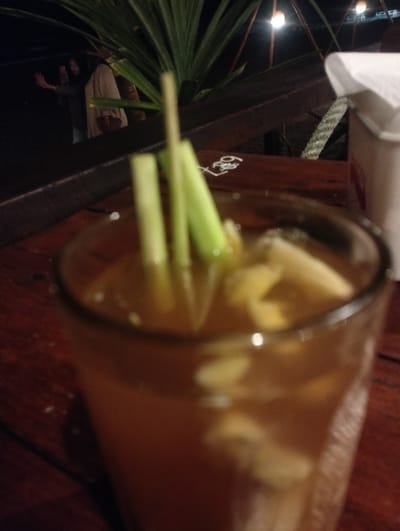 The image displays a close-up, slightly out-of-focus shot of a glass containing a light brown beverage, possibly a tea or herbal drink, situated on a dark wooden table. Inside the glass, several light-colored, irregular pieces, resembling ginger or cut fruit, are visible, along with two slender green stalks, likely lemongrass, standing upright as a garnish or ingredient.
To the right of the glass, on the same wooden table, a white paper napkin dispenser or cup is partially visible. Further back on the table surface, some faint white markings, including the number "28" and other indistinct symbols, are visible in one of the detailed crops.
The background is predominantly dark and blurry, indicating a nighttime setting or a dimly lit indoor environment. A large, leafy green plant, possibly a palm or similar tropical foliage, occupies the upper left portion of the background. Beyond the plant, a person is faintly discernible in the distance, appearing to hold a device or gesture with an arm. Several small, glowing artificial lights, resembling bare bulbs or decorative lights, can be seen in the far background, suggesting an overhead structure or canopy. The scene evokes a casual evening atmosphere, likely at an outdoor or semi-outdoor cafe or restaurant in Keudekruenggeukueh, Indonesia. The primary focus is on the prepared drink.