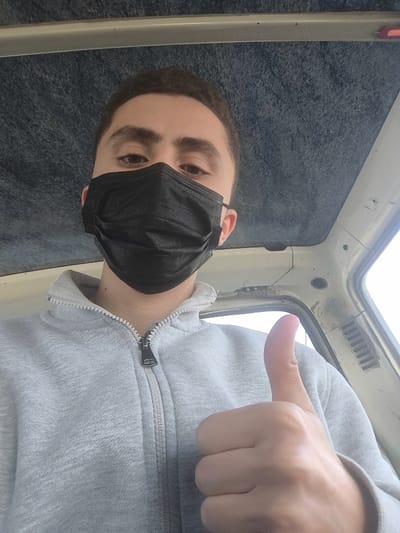 A young male individual, appearing to be in his late teens or early twenties, is depicted from a low-angle perspective, seated inside a vehicle in Dahiyat Harasta, Syria. He has short dark hair and dark eyes, partially visible above a black disposable-style face mask that covers his nose and mouth. He is wearing a light grey full-zip sweatshirt with a metallic zipper pull. His right hand is raised, forming a thumbs-up gesture with the thumb pointed upwards. The vehicle's interior features a dark blue/grey textured headliner and a light-colored window frame or pillar on the right side of the frame, showing an indistinct bright outdoor light through the window.