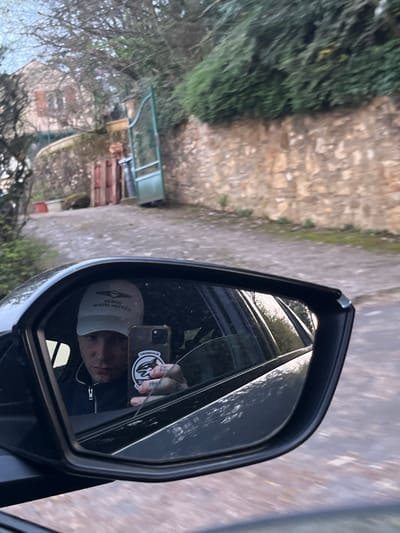 The image displays a reflection in a vehicle's passenger-side rearview mirror, capturing a male individual actively taking a selfie. The man wears a light-colored baseball cap with the mirrored text "GENESIS SCOTTISH OPEN" and a dark, zippered jacket. He holds a dark-colored smartphone, prominently displaying a black and white orca logo sticker on its back, with tattoos visible on his fingers. A light-colored mark or tattoo is present on his left cheek (viewer's right).
The scene outside the vehicle, located in Gif-sur-Yvette, France, features an uneven, possibly paved or gravel pathway sloping gently uphill. To the right of the pathway, a substantial stone wall is visible, capped with a dense green hedge. Further along the right, an open green metal gate stands. On the left side of the path, a pair of reddish-brown wooden gates are partially open, leading towards a light-colored stone or stucco house with terracotta-colored window shutters, partially obscured by trees with visible bare branches. The lighting indicates daylight conditions.