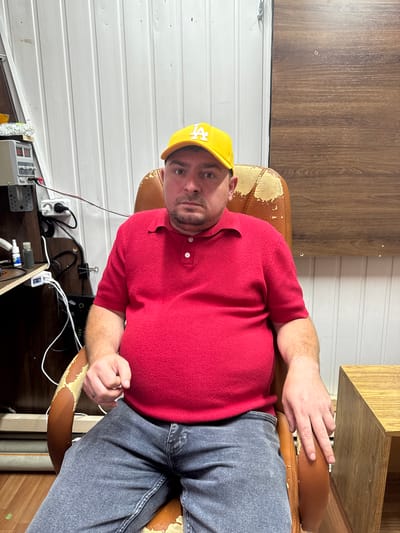 A man wearing a yellow cap and a red polo shirt is sitting in a worn leather office chair. He is looking directly at the camera with a neutral expression. The background consists of a white, corrugated metal wall and a wooden panel. To the left of the man, there is a desk with electronic equipment and cables. To the right, a portion of a wooden table is visible. The overall scene appears to be an indoor workspace or utility area.