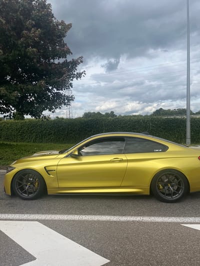 A bright yellow two-door BMW M4 coupe is stationary, positioned in profile view, facing left to right, on an asphalt surface in Dorlisheim, France. The vehicle features dark gray multi-spoke alloy wheels with visible BBS branding on the center caps and yellow brake calipers, also marked with BBS. The car's roof is dark in color, and its windows are tinted. A small white sticker with "@dri_s4" is present on the rear side window. White painted road markings, resembling an arrow or chevron pattern, are visible on the asphalt in the foreground.

The environment under a heavily overcast, dark gray sky. To the left, a large deciduous tree with dark green foliage partially obscures a metallic lattice-style utility pylon. A dense, green, manicured hedge forms a horizontal barrier across the mid-ground. Above the hedge, multiple horizontal power lines traverse the sky. A slender, silver utility pole stands on the far right. Distant trees and a faint elevated structure are visible beyond the hedge. No individuals are present in the scene.