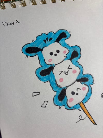 The image is a hand-drawn illustration on a piece of paper, likely from a notebook given the spiral binding visible at the top. The drawing depicts three cartoon dog-like characters stacked vertically on a stick, resembling a skewer of treats. The characters are white with black ears and eyes, and pink cheeks. The top character has simple dots for eyes and a neutral expression. The middle character has its tongue sticking out and its eyes closed, conveying a playful or excited mood. The bottom character has its eyes closed in a smile. The characters are outlined in black and filled with blue, giving them a fluffy, cloud-like appearance around their heads and bodies. The stick is colored orange. To the left of the skewer, there are two small, detached, rectangular shapes outlined in black. Above the drawing, in the upper left corner, the words "Day 1." are written in black ink. The background is a plain white surface. There is no indication of the city of Velbert, Germany, in the image.