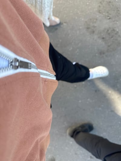 The image is a close-up, angled shot of two individuals on a paved surface, likely a sidewalk or street, in the city of Kara-Suu, Kyrgyzstan. The foreground is dominated by the textured fabric of a rust-colored garment, possibly a hoodie or jacket, with a prominent silver zipper. The zipper's teeth and pull tab are clearly visible, suggesting the garment is partially open. To the right of the zipper, another person's legs are visible, wearing black pants and white perforated shoes. The ground beneath them is a light gray, speckled asphalt with some debris. In the background, further down the paved surface, another person's legs are seen wearing dark pants and dark shoes. The lighting suggests it is daytime, with a bright, possibly sunlit area casting a shadow on the ground to the right.
