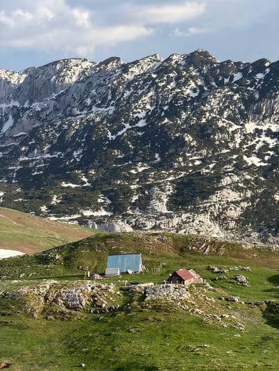 The image depicts a rugged mountain landscape, likely in the vicinity of Komarnica, Montenegro, though specific location is difficult to ascertain without more context. The left portion of the image is dominated by a steep, rocky mountain face with patches of snow and sparse vegetation clinging to its sides. To the right, the terrain transitions to a grassy slope interspersed with rocks and two small buildings. One building has a rusty red roof, and the other has a striped blue roof. The sky is visible in the upper left corner.