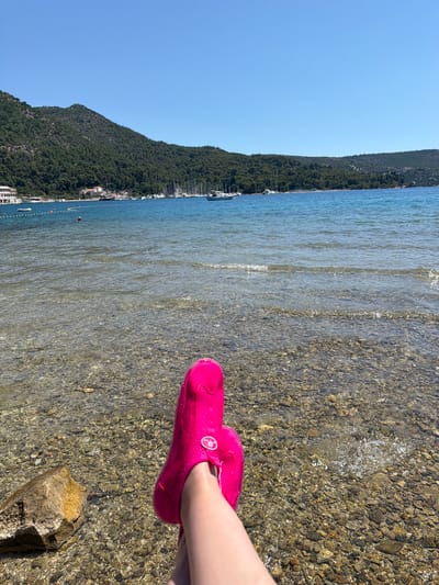 A person's feet, wearing bright pink water shoes, are visible in the foreground, resting on a rocky beach at the edge of clear, shallow water. The water is calm with gentle ripples, and the seabed is composed of small, multi-colored stones. Beyond the shoreline, a lush, green, tree-covered mountain slopes down towards the sea. A line of buildings and boats is nestled along the coast at the base of the mountain, suggesting a small town or village. The sea stretches out to the horizon, meeting a clear, bright blue sky. The sun appears to be high in the sky, casting shadows and illuminating the scene with bright light, indicating it is likely midday on a sunny day. The overall impression is a peaceful and idyllic coastal scene, likely a vacation destination. No visible text is present in the image.