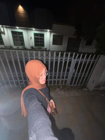 A young woman wearing a brown hijab and glasses is taking a selfie at night in front of a white building with green-framed windows. She has a dark sweater and a brown bag slung across her shoulder. The building is partially illuminated by a light from above. A metal fence runs along the front of the property. The scene appears to be a residential area in Tanggul, Indonesia.