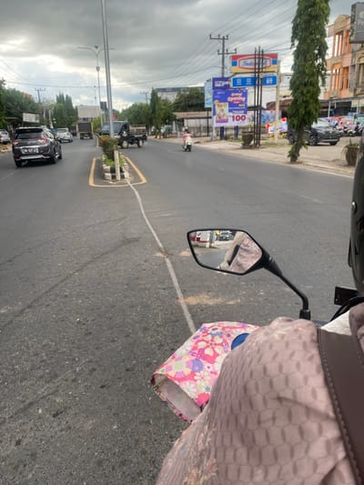 The image captures a street scene in Sungai Paring, Indonesia, under an overcast sky. The perspective is from a motorcycle, looking forward down a two-lane road. On the left side of the road, several cars are visible, including a dark SUV with license plate "DA 1417 AJ" in the foreground. Further down the road, other vehicles, including trucks and a white car, are also present.
The right side of the road features a sidewalk area with several billboards. One prominent billboard displays the "Indomaret" logo. Another billboard announces a "Grand Opening" for "Me-O" with promotional pricing. Trees line the sidewalk, and buildings with orange and light-colored facades are visible in the background. A few motorcycles are parked on the sidewalk.
A person is riding the motorcycle from which the photo is taken. Their arm and shoulder are visible in the lower right corner, wearing a light brown patterned garment. A patterned bag is also visible on the motorcycle. The motorcycle's side mirror reflects a black car driving on the road. The road itself appears to be asphalt with white lane markings. The overall environment suggests a moderately busy urban or suburban area.