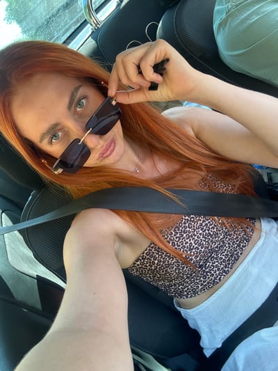 A woman with long, auburn hair is lying down in a car, her head tilted back and resting on the seat. She is wearing sunglasses with dark lenses and a gold frame, which she is holding up to adjust. Her top is a white tank top layered over a leopard-print bandeau. A black seatbelt crosses over her chest. Her eyes are light green and wide open, and her lips are slightly parted. She is wearing a delicate silver necklace with a small pendant. Her left hand is holding a small, black object, possibly car keys. The car appears to be parked, with the passenger seat visible and a glimpse of a window showing greenery and sunlight, suggesting it is daytime and the weather is clear. The scene conveys a relaxed, possibly candid moment.