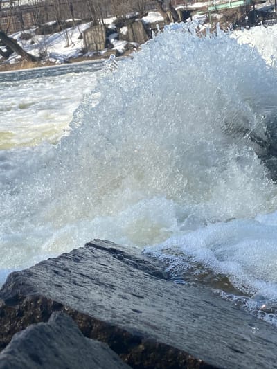 The image depicts a close-up of a waterfall crashing against dark, rough rocks. The water appears turbulent and frothy. In the background, beyond the waterfall, there's a glimpse of trees and snow-covered ground. The location is Postol, Russia. There are no people in the image.