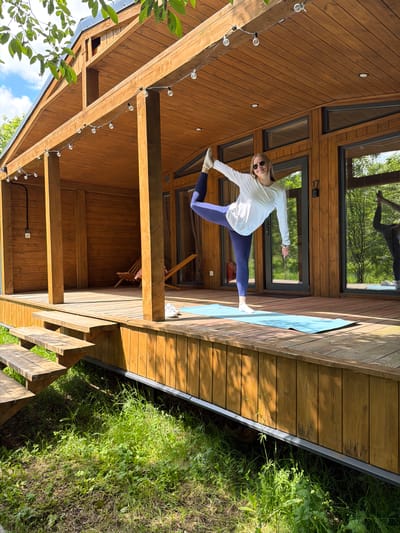 A woman is captured performing a dynamic yoga or dance pose on a wooden deck next to a modern wooden house, set in Vozhoiskii, Russia. She appears to be in her 20s or 30s, dressed in a white long-sleeved top, dark blue leggings, and white socks, with sunglasses perched on her head. Her pose involves balancing on one leg with the other extended backward and upward, while her arms are outstretched for balance, suggesting a warrior III or similar balancing posture.

She is standing on a light blue yoga mat laid out on the spacious wooden deck. The deck is attached to a contemporary wooden house or cabin, characterized by its natural wood paneling and large, angular windows that reflect the lush green trees and clear blue sky. The design hints at a modern, nature-integrated living space. String lights are visible, draped along the wooden beams of the deck's overhead structure.

The surrounding environment is rich with greenery, featuring dense grass and foliage visible next to and below the deck, indicating a natural, possibly rural or forested setting. The scene is illuminated by bright daylight, suggesting mid-morning or afternoon, under a clear sky. The overall impression is one of active well-being and tranquility in a beautiful, naturalistic environment. No visible text is present in the image.