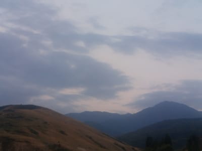 The image depicts a mountainous landscape, likely taken during twilight or overcast conditions given the muted colors and soft light. The foreground features a sloping, grassy hillside, rendered in shades of brown and gold, suggesting dry vegetation. The midground is dominated by the hazy, blue silhouettes of successive mountain ranges. In the background, the sky is a pale gray, filled with soft, diffuse clouds. The overall impression is one of natural, undeveloped terrain. No people, specific structures, or identifiable landmarks are visible. The scene is consistent with the mountainous geography of Jardy-Su, Kyrgyzstan, known for its rolling hills and expansive mountain vistas.