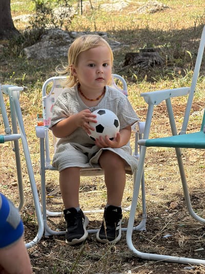 In an outdoor setting, a fair-skinned toddler with blonde hair sits in a highchair, holding a small, black-and-white soccer ball. The child is wearing a light-colored, loose-fitting outfit and dark sneakers with white stripes. A brown beaded necklace adorns their neck. The highchair is white with a light blue and green support bar. The ground is covered in dry grass and leaves. In the bottom right corner, part of a person's leg wearing blue shorts is visible. The location is suggested to be in the Zvulun Regional Council, Israel.