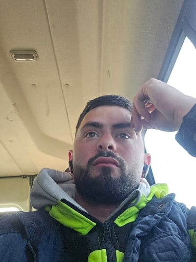 An adult male is depicted in a low-angle, headshot-style photograph taken inside a vehicle, the location of which is specified as Opalchenets, Bulgaria. The subject has short dark hair, a full dark beard, and dark eyebrows, and is looking upwards with his mouth slightly ajar. He is wearing black wireless earbuds in both ears. His right hand, with fingers bent, is positioned near his right temple. He is dressed in layered clothing, consisting of a grey hooded sweatshirt with a visible white collar lining, worn under a dark-colored jacket featuring neon yellow/green zipper placket and collar accents. The vehicle's interior includes a light-colored (beige/tan) headliner with a rectangular overhead light fixture visible in the upper left, and a bright, out-of-focus window on the right side of the frame, indicating external light. The headliner shows minor surface imperfections and possible faint discoloration. No other individuals are visible.