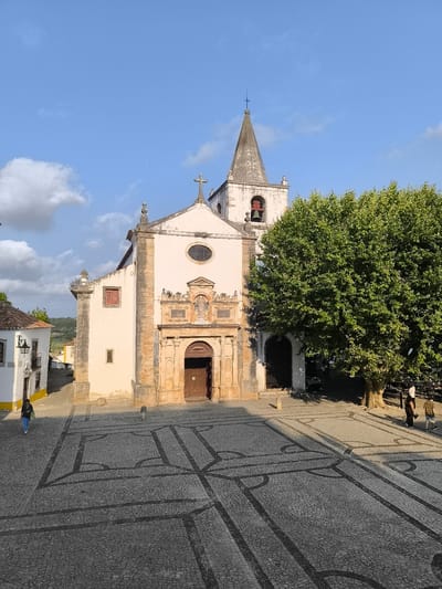 This image depicts the main square of Óbidos, Portugal, featuring the Church of Santa Maria. The church is a white building with a prominent bell tower topped by a spire and cross. Its facade is adorned with stonework and a large wooden door. To the left of the church, a smaller building with white walls and a red-tiled roof is visible, characteristic of the town's architecture. The square itself is paved with traditional Portuguese cobblestones, featuring a distinct black and white mosaic pattern. A large, mature tree stands to the right of the church, casting shadows across the square. Two individuals are walking across the square, one in the foreground to the left and two others in the background to the right. The sky above is a clear blue with scattered white clouds. The scene is bathed in natural sunlight, suggesting it was taken during the daytime.