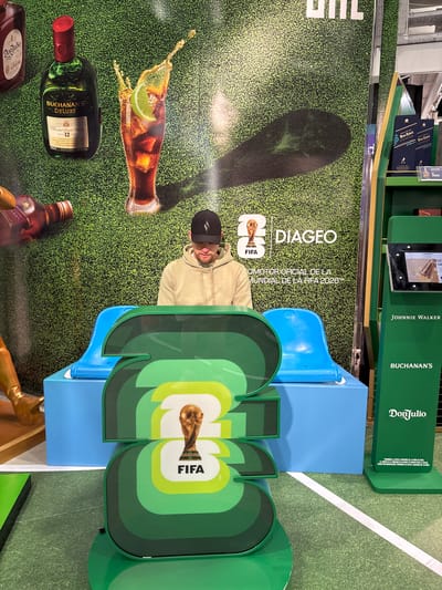 This media file shows a display for Diageo brands, specifically Buchanan's and Don Julio, in collaboration with FIFA for the 2026 World Cup. The display features a large backdrop with a grass texture, a stylized image of a drink with lime and ice, and bottles of Buchanan's Deluxe Scotch Whisky. In the foreground, a man wearing a black baseball cap is partially visible, looking towards the display.
A prominent element of the display is a large green cutout with the FIFA World Cup trophy and the FIFA logo. There are also blue structures that seem to be part of the display, and a green display tower behind the backdrop with bottles of Johnnie Walker Blue Label and text that appears to be promotional. The overall setting suggests a retail or duty-free environment, likely an airport given the mention of Ezeiza, Argentina.
The scene is well-lit, indicating it's likely during daytime. The focus is on the brand promotion, with no specific activity or emotion being conveyed beyond that. The visible text includes brand names, product details like "AGED 12 YEARS," and promotional messages in Spanish, such as "PROHIBIDA LA VENTA A MENORES DE 18 AÑOS DE EDAD" (Sale to minors under 18 years of age is prohibited). The presence of the FIFA branding suggests this is a timely promotion related to the upcoming World Cup.