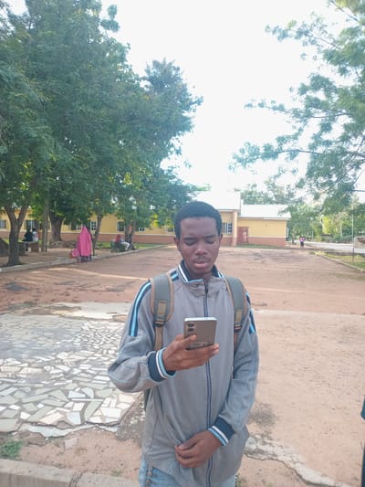 In Girei, Nigeria, a young man stands outdoors, facing the camera and looking down at his phone. He is wearing a gray zippered jacket with blue and black stripes on the sleeves and collar, and a backpack with green and brown straps. The ground in front of him is a mix of sand and broken tile mosaic. In the background, there is a yellow building with white trim and windows, surrounded by trees and other people. One person in the background is wearing a bright pink headscarf. The sky is bright and overcast.