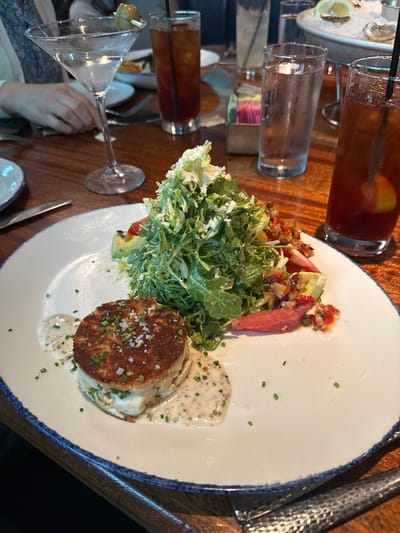 The image shows a plate of food on a wooden table, likely in a restaurant. The plate contains a crab cake with a creamy sauce, a salad with watermelon and greens, and a garnish of herbs. To the left of the plate, there is a martini glass with an olive and other beverages. The presence of such dishes suggests a fine dining experience. Given the specification of Southlake, United States of America, it is probable that the restaurant is located in Southlake. The environment suggests an indoor setting with good lighting.