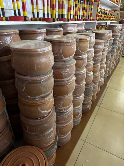 This is a medium-shot, eye-level, outdoor photograph taken in the city of Acıpayam, Turkey. The image captures a detailed view of stacks of terracotta pots, some wrapped in plastic, arranged on shelves and the floor of a marketplace or shop. The pots are various sizes and shades of brown. In the background, the shelves are also stocked with what appear to be wooden objects with red and yellow bands, possibly decorative items or tools. The floor is tiled in a light beige color. The lighting suggests it is daytime, with natural light illuminating the scene.