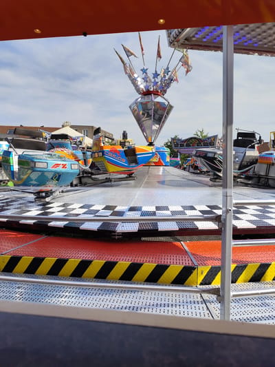 The image displays a section of a fairground ride, likely a spinning amusement ride with multiple car-like pods designed to resemble race cars. The pods are brightly colored in blue, orange, and yellow, with some featuring logos like "F1" and "Red Bull." The ride platform is covered in a checkered black and white pattern. The ride is situated outdoors, with a partly cloudy blue sky visible in the background. In the distance, buildings with tiled roofs, characteristic of residential areas in the Netherlands, can be seen. The overall impression is that of a typical funfair attraction. The location is identified as Dongen, Netherlands, though there is no specific landmark or context in the image to confirm this definitively. There are no people visible in the image, and the ride appears to be stationary. The image is taken from a low-angle perspective, focusing on the ride's structure and decorative elements.