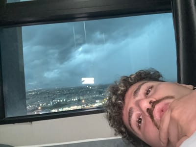 The media file is a selfie taken by a person with curly hair and a mustache. The person is looking towards the camera with a neutral expression. The background shows a window with a view of a city at night, with lights visible in the distance. The sky appears cloudy and dark, suggesting it might be evening or night. The scene is indoors, likely in a room with a window overlooking the city. The person's face is partially obscured by their hand. There is no visible text in the image. The rotation of the image suggests it was taken by holding the camera sideways.