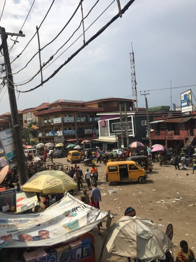 The image depicts a crowded, unpaved commercial street or market intersection in Ojo, Nigeria, under a partly cloudy sky. The scene is characterized by dense human activity, numerous vehicles, and varied commercial structures.
In the foreground, a prominent wooden utility pole supports a complex tangle of electrical wires, many appearing unsecured. Below, several market stalls are established, featuring assorted umbrellas and tarpaulin canopies, one yellow umbrella displaying "MTN Data Bundles." Merchandise, including electronic devices, is visible on tables under these covers. The unpaved ground surface is uneven, dusty, and littered with debris, including plastic bags and paper.
The mid-ground reveals a bustling confluence of pedestrians and vehicles. Numerous yellow commercial minibuses are parked or navigating the unpaved area, some with open doors. Other vehicles include cars and auto-rickshaws. A large number of individuals, dressed in various casual attire, are walking, standing, or interacting within the crowded space. Many market vendors operate under assorted colorful umbrellas.
In the background, a mix of commercial buildings is visible. Multi-story structures with orange-brown roofs dominate the left and center, featuring numerous windows and signs on their facades. A more modern, multi-level building with a grey and purple facade stands prominently in the mid-right. To its right, older, single-story structures with corrugated metal roofs are present. A tall, red and white lattice communication tower is visible in the distant background, along with additional utility poles and overhead lines. Several billboards and commercial signs are affixed to poles and buildings throughout the scene.
