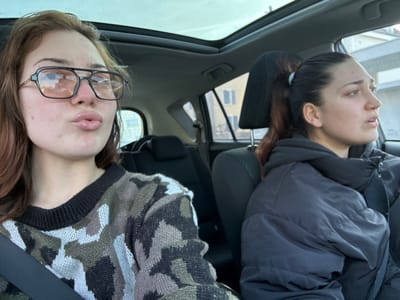 The scene depicts the interior of a passenger vehicle, likely in motion or recently stopped, in Souffelweyersheim, France. Two young adult females occupy the front seats.
The female in the front passenger seat, positioned on the left, has long, reddish-brown hair and wears large, dark-framed aviator-style eyeglasses with orange-tinted lenses. She is attired in a camouflage-patterned sweater composed of dark green, black, and light gray shapes. Her head is angled slightly towards the camera, and her lips are pursed into a distinct "kissing" or "pout" expression. A black seatbelt crosses her left shoulder and chest.
The female in the driver's seat, positioned on the right, has dark hair pulled back into a high ponytail secured with a light-colored hair elastic. She wears a dark, possibly black, puffy winter jacket. Her face is in profile, oriented forward and to the right, consistent with operating a vehicle. Her mouth is slightly open, and her gaze is directed outwards.
The vehicle's interior features dark upholstery and a panoramic sunroof, through which a bright, ambient sky is visible. Outside the windows, blurred architectural structures with discernible window frames are present, indicating an urban or suburban environment. The passenger appears to be posing for a photograph, while the driver maintains focus on the external surroundings.