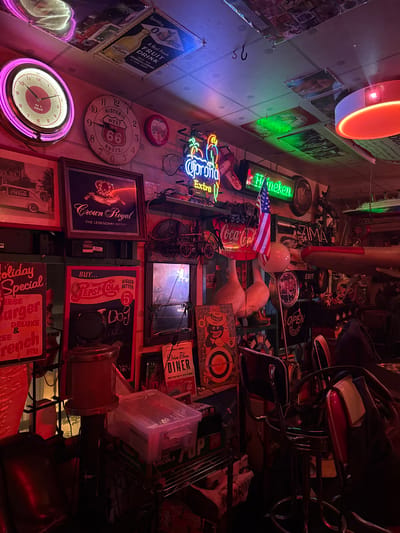 This media file captures a dimly lit, cluttered interior, likely a bar or diner, filled with retro-themed memorabilia. Neon signs for brands like "Heineken," "Corona Extra," and "Coca-Cola" illuminate the space, casting colorful glows on the walls. Various vintage objects are displayed, including clocks, framed advertisements for soda and food, and what appear to be bar stools. A US flag is also visible, adding to the nostalgic American diner aesthetic. The overall impression is one of a vibrant, albeit crowded, establishment steeped in a mid-20th century American pop culture theme. The text visible includes brand names, slogans like "BIGGER BETTER," "A Real Fresh FRUIT DRINK," and "THE LEGENDARY IMPORT," as well as directional signage like "EXIT" and "OPEN." There are no visible people in the frame, suggesting the photograph was taken during off-hours or the subjects are out of frame. The lighting suggests it is nighttime or the interior is intentionally kept dark to enhance the neon signs.
