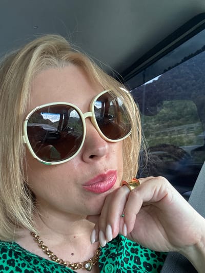 A close-up, selfie-style shot captures a fair-skinned woman with blonde hair and light skin tone, wearing large, cream-rimmed sunglasses with brown lenses. Her lips are a bright pink, and she is wearing a gold necklace with a heart-shaped pendant and a large, abstract gold ring on her right hand, which is resting on her chin. She is also wearing a green top with a black leopard print. The background appears to be the interior of a vehicle, with reflections of a road and mountains visible in the sunglasses. The scene is set in the city of Kepsha, Russia.