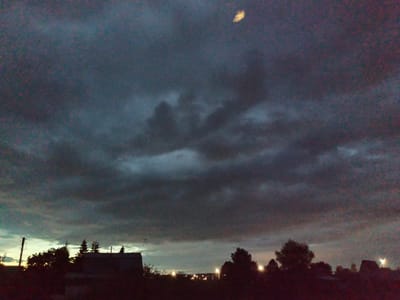 This image depicts a low-light, possibly dusk or night scene in Осоргино, Russia. The upper two-thirds of the frame are dominated by a heavily clouded sky, featuring dense, dark gray and bluish-gray clouds with varying textures and opacities. In the upper-central portion, a distinct, bright, elongated, yellowish light source is visible against the dark cloud cover, indicative of a celestial body. The lower third of the image presents a dark silhouette of a landscape. This silhouette is composed of numerous trees, appearing both coniferous and deciduous, which form an irregular treeline across the horizon. Towards the left of the frame, a building with a visible roofline and a utility pole are clearly silhouetted. Multiple distant artificial light sources, consistent with streetlights or lights from structures, punctuate the dark mid-ground horizon, primarily on the right side, suggesting a populated area. The overall image exhibits noticeable photographic noise, consistent with low-light conditions. No individuals or specific actions are discernible within the frame.