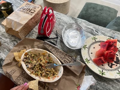 This is a forensic, matter-of-fact description of an image taken on a granite countertop. The scene depicts remnants of a meal, suggesting a recent dining experience.
In the foreground, a white bowl contains a partially eaten mixture of what appears to be rice, beans, corn, and pico de gallo. Several tortilla chips are scattered around the bowl and on a brown paper bag. A silver fork rests inside the bowl. To the left, a hand with pink painted fingernails is holding a tortilla chip.
Further to the left, a brown paper bag with "HANDCRAFTED CHIPS MADE FRESH EVERY DAY" printed on it is partially open. A dark green glass container with what looks like ice and possibly a drink is next to the chip bag.
To the right of the food bowl, an empty aluminum foil container sits on the countertop. Next to it is a decorative plate with several slices of watermelon and scattered watermelon seeds.
In the background, a small, bright red patent leather purse with a zipper and white trim is visible. A pair of black sunglasses is also on the countertop. Two large, dark green cushions are propped up in the background, suggesting a casual seating area. The overall environment appears to be indoors, on a kitchen or dining surface.
The image does not contain specific visual cues to pinpoint its exact location within Jacksonville Beach, United States of America. However, the presence of food items commonly associated with fast-casual dining establishments, such as the Chipotle-branded paper bag, suggests a meal consumed from such a restaurant. The overall setting is consistent with a residential or dining area in a coastal city like Jacksonville Beach.